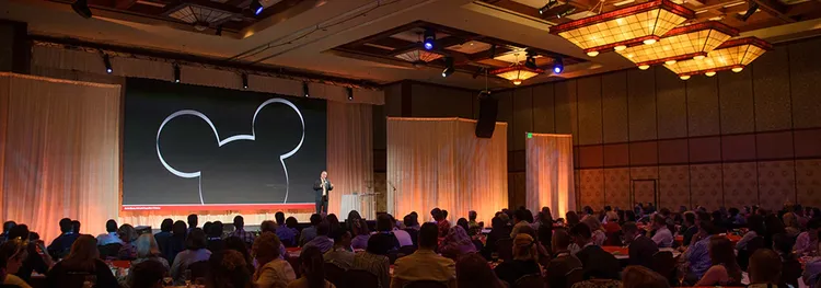 Attendees sitting at tables listening to a speaker in a conference room that has a large Mickey Mouse design on the wall