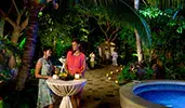 Two women with drinks talk while standing at an outdoor cocktail table near a candlelit stone path