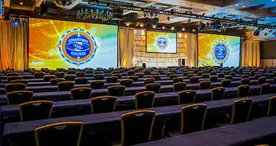 A ballroom set up with rows of seats, a stage and 2 large screens for a conference presentation