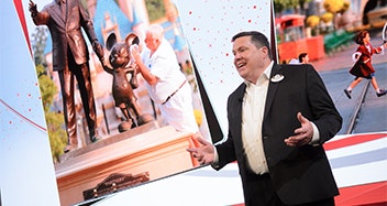 A man in a suit giving a speech next to a life size image of the Partners statue in front of Cinderella Castle