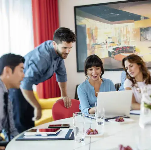 A group of men and women in professional attire look at the screen of a laptop in a meeting room