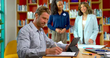 A man working at a table with a tablet and pad of paper while 2 women stand at the opposite end of the table