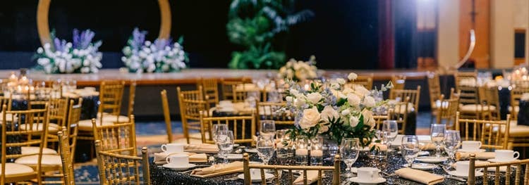 A ballroom with round tables, chairs, table settings, flower arrangements and a stage decorated with a Mickey Mouse shaped arch