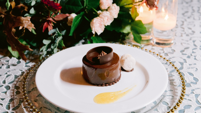 An elegantly plated chocolate dessert with a caramel sauce alongside candles and flowers