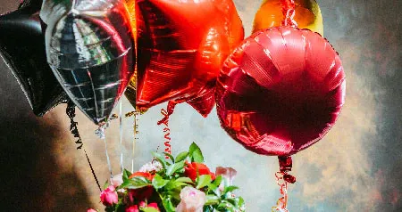Several Mickey Mouse themed balloons, a tray of 6 cupcakes and a vase of flowers sitting on a table